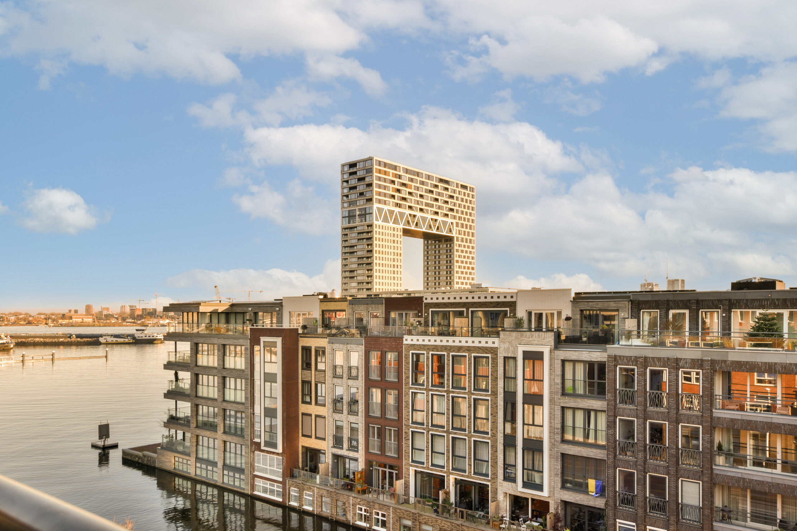 Moderne appartementencomplexen met een iconisch hoogbouwproject op de achtergrond, gelegen aan het water, onder een heldere lucht.