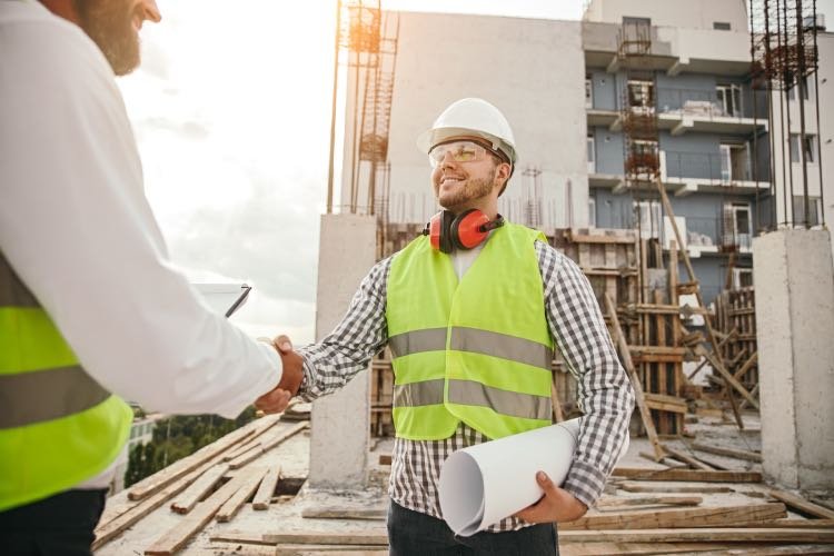 Een bouwprofessional in veiligheidskleding schudt de hand van een klant op een actieve bouwplaats, met zonlicht op de achtergrond.