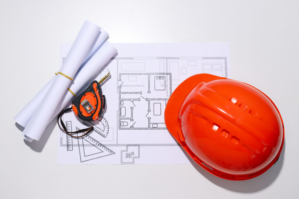 Bouwtekeningen, een meetlint en een oranje veiligheidshelm op een werktafel, symbool voor bouwprojecten en expertise.